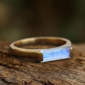 Puede incluir: Un anillo de oro con una piedra preciosa rectangular de color azul claro. El anillo está colocado sobre un trozo de madera marrón oscuro, con un fondo verde borroso. La piedra preciosa tiene un brillo sutil e iridiscente, y la banda del anillo parece ser de oro cepillado.