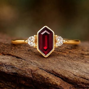 May include: A gold ring featuring a central, elongated, hexagonal-cut, deep red gemstone. Two smaller, round, clear gemstones flank the central stone. The ring is set on a wooden surface.