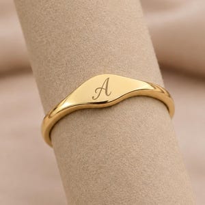 Könnte beinhalten: Ein goldfarbener Siegelring mit einer glatten, polierten Oberfläche und dem eingravierten Großbuchstaben A in Schreibschrift auf der flachen Oberseite. Der Ring ist schmal und weist einen gleichmäßigen Umfang auf.