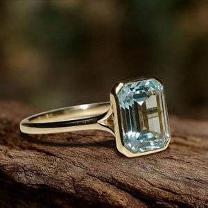 May include: A gold ring with a large, rectangular, pale blue gemstone. The gemstone is set in a gold bezel, and the band is polished gold. The ring is shown on a wooden surface.