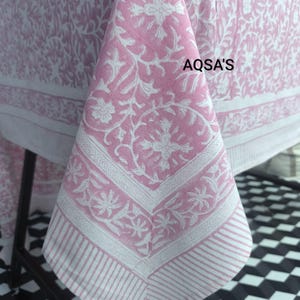 Puede incluir: Un mantel rosa y blanco con estampado floral y borde detallado. El mantel presenta un patrón floral repetido y el texto "AQSA'S" en la esquina superior derecha. El borde tiene un diseño a rayas.