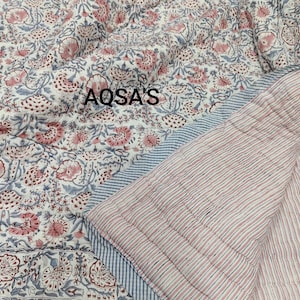May include: A quilted textile featuring a white background with a repeating floral pattern in muted pink and blue tones. The text AQSA'S is printed on the fabric. The reverse side displays a pattern of thin vertical pink and blue stripes.
