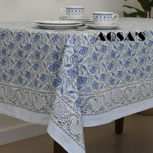 Può includere: Una tovaglia rettangolare con base bianca, un motivo ripetuto di tralci floreali blu e verde salvia e un bordo a righe. Il testo AQSA'S è stampato in nero sul tessuto. Due tazze e piattini in ceramica bianca sono posizionati sul tavolo.