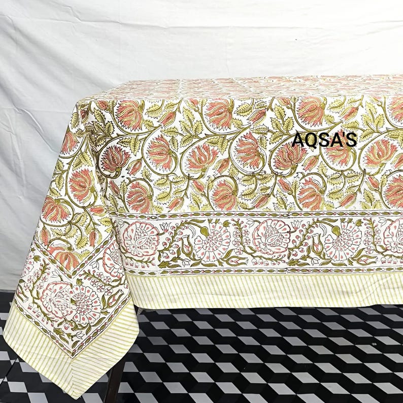 K&ouml;nnte beinhalten: Eine Tischdecke mit Blumenmuster mit wei&szlig;em Hintergrund und einem sich wiederholenden Muster aus rosa und gr&uuml;nen Blumen und Bl&auml;ttern. Der Rand weist ein detailliertes Blumendesign auf. Der Text "AQSA'S" ist auf der Tischdecke zu sehen.