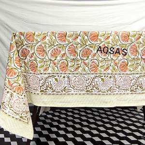 K&ouml;nnte beinhalten: Eine Tischdecke mit Blumenmuster mit wei&szlig;em Hintergrund und einem sich wiederholenden Muster aus orangefarbenen und gr&uuml;nen Blumen. Der Rand hat ein Blumendesign mit rosa Bl&uuml;ten. Der Text "AQSA'S" ist auf die Tischdecke gedruckt.