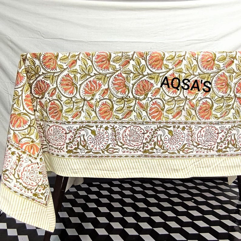 K&ouml;nnte beinhalten: Eine Tischdecke mit Blumenmuster mit wei&szlig;em Hintergrund und einem sich wiederholenden Muster aus orangefarbenen und gr&uuml;nen Blumen. Der Rand hat ein Blumendesign mit rosa Bl&uuml;ten. Der Text "AQSA'S" ist auf die Tischdecke gedruckt.