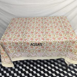K&ouml;nnte beinhalten: Ein Tischtuch mit Blumenmuster mit einem sich wiederholenden Muster aus rosa Blumen und gr&uuml;nen Bl&auml;ttern auf wei&szlig;em Grund. Das Tischtuch hat einen cremefarbenen Rand mit floralem Design und dem Text "AQSA'S".