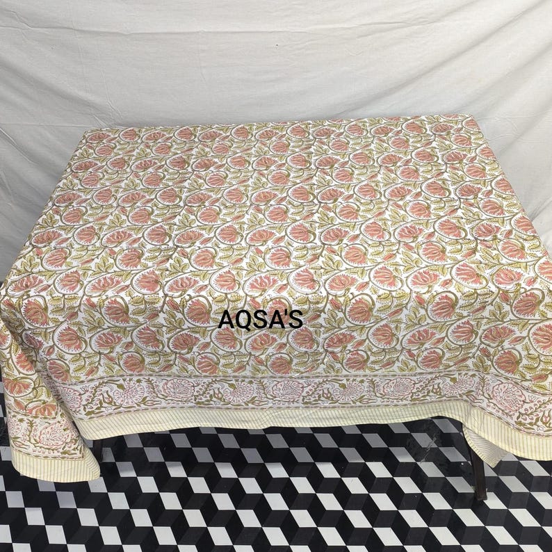 K&ouml;nnte beinhalten: Ein Tischtuch mit Blumenmuster mit einem sich wiederholenden Muster aus rosa Blumen und gr&uuml;nen Bl&auml;ttern auf wei&szlig;em Grund. Das Tischtuch hat einen cremefarbenen Rand mit floralem Design und dem Text "AQSA'S".