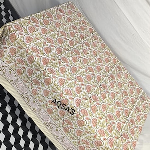 K&ouml;nnte beinhalten: Gefalteter Stoff mit einem sich wiederholenden Blumenmuster in Rosa, Gr&uuml;n und Wei&szlig;. Der Stoff hat einen floralen Rand und der Text "AQSA'S" ist sichtbar. Dieser Stoff ist wahrscheinlich f&uuml;r Textilien oder Heimdekoration bestimmt.
