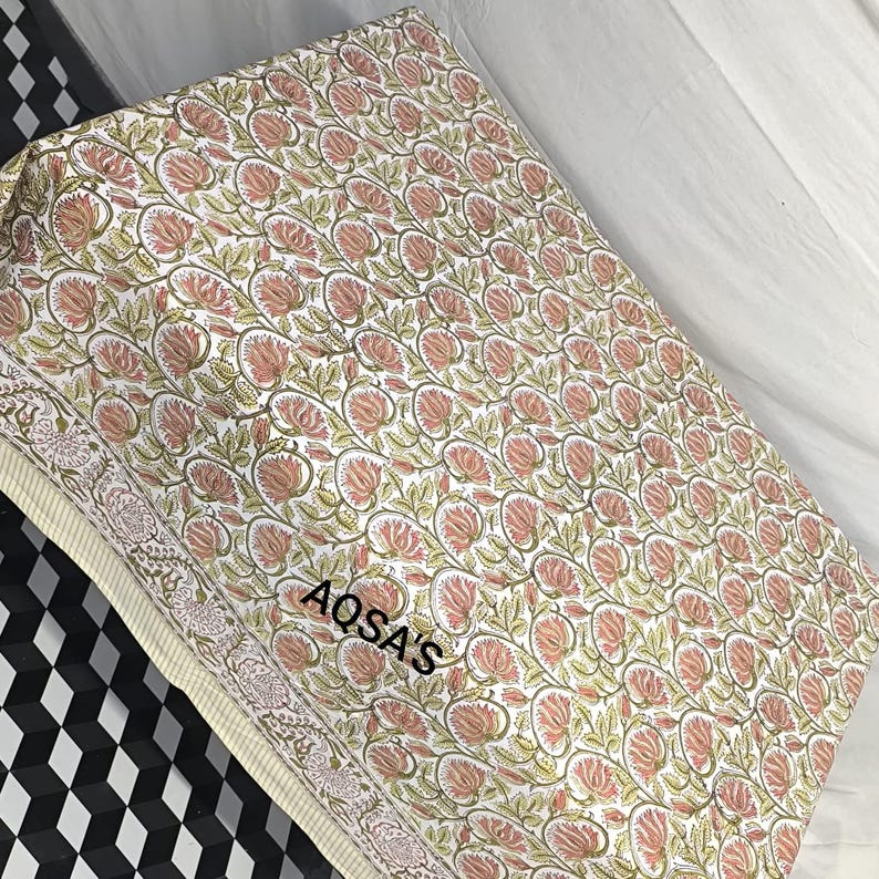 K&ouml;nnte beinhalten: Gefalteter Stoff mit einem sich wiederholenden Blumenmuster in Rosa, Gr&uuml;n und Wei&szlig;. Der Stoff hat einen floralen Rand und der Text "AQSA'S" ist sichtbar. Dieser Stoff ist wahrscheinlich f&uuml;r Textilien oder Heimdekoration bestimmt.