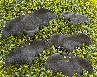 May include: Four dark gray felt bat ornaments of varying sizes, each featuring two small yellow circular eyes and a stitched yellow mouth. The bats have scalloped wing edges and are attached to thin, clear hanging strings for display.