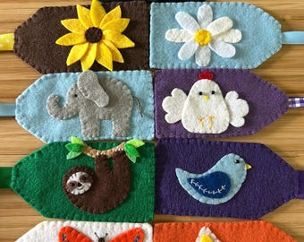 May include: Eight felt fabric tags with different embroidered designs. The designs include a mushroom, sunflower, daisy, elephant, chicken, sloth, bird, butterfly, and star.