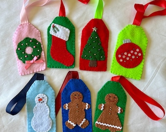 May include: A collection of handmade felt Christmas gift tags in various colors. Each tag features a festive design, including a wreath, stocking, Christmas tree, ornament, snowman, and gingerbread people. They have colorful ribbon loops for hanging.
