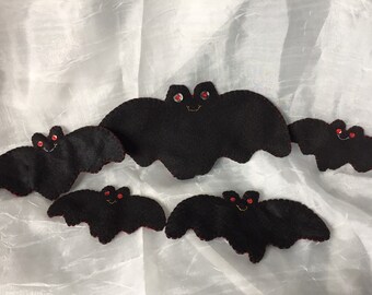May include: A collection of six black felt bat decorations with red gem eyes and red stitching around the edges. Each bat has a smiling face embroidered with yellow thread. The bats are arranged on a white, textured background.