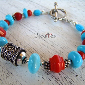 May include: A turquoise and red beaded bracelet with a silver toggle clasp and a silver bead with an intricate design. The bracelet is on a white wooden surface.