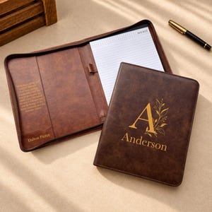 Puede incluir: Una carpeta de cuero marrón con un bloc de notas en el interior. La cubierta presenta una letra A dorada, detalles florales y el nombre Anderson. El panel interior izquierdo incluye una cita impresa y el nombre Dalton Pierce. Un bolígrafo negro está cerca.
