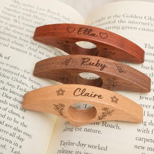 May include: Three curved wooden page holders with a central hole for a thumb. Each piece is engraved with a name: Ella with hearts, Ruby with birds, and Claire with stars. All three feature delicate floral line art engravings on the wood surface.