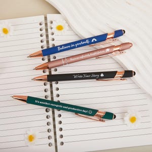 Puede incluir: Cuatro bolígrafos con detalles en oro rosa y citas inspiradoras sobre un cuaderno rayado. Los bolígrafos son azules, rosas, negros y verdes. Las citas incluyen "Believe in yourself," "Have an infinitely bright future," "Write Your Story," y "It's another energetic and productive day!"