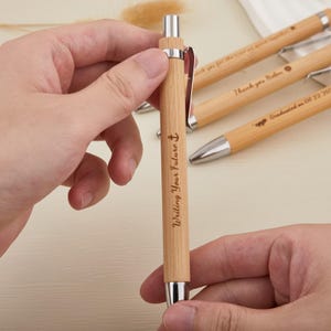 Op de afbeelding: Een lichtbruine bamboe pen met zilveren details en een zilveren clip. De pen is gegraveerd met de woorden "Writing Your Future" en een ankersymbool. Andere pennen met gegraveerde tekst staan op de achtergrond.