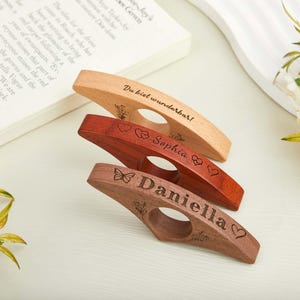 May include: Three wooden book page holders in varying shades of brown. The top one is light, the middle is reddish-brown, and the bottom is dark brown. Each has a circular opening and engraved text and designs. The top one says "Du bist wunderbar!"
