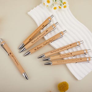 Peut inclure: Plusieurs stylos en bambou avec des détails argentés. Chaque stylo porte une inscription personnalisée, comme "Every day is a gift" et "Merci pour votre soutien." Ils sont disposés sur une surface blanche texturée, avec des fleurs en arrière-plan.