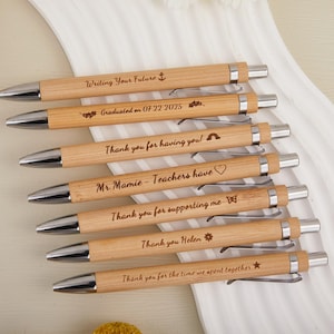 Op de afbeelding: Een verzameling gepersonaliseerde houten pennen met zilveren accenten. Elke pen is gegraveerd met een unieke boodschap, waaronder zinnen als "Writing Your Future" en "Thank you for having you!". De pennen liggen op een witte ondergrond.