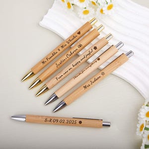 Peut inclure: Plusieurs stylos en bambou personnalisés. Les stylos ont des pointes dorées ou argentées et sont gravés de noms, de dates et de phrases telles que "Mr. Franklin & Mrs. Wheeler" et "S.R 09.02.2025". Les stylos sont de couleur brun clair.