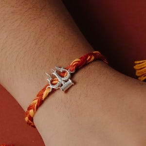 Puede incluir: Una pulsera trenzada roja y amarilla con un dije plateado en forma de tridente. La pulsera está en una muñeca sobre un fondo marrón rojizo. También se ven hebras trenzadas adicionales de color rojo y amarillo.