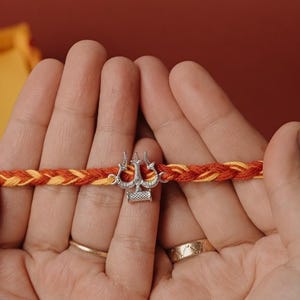 May include: A braided bracelet with red and yellow threads, featuring a silver-colored charm in the shape of a trident. The bracelet is held in open hands against a red background. A yellow box with the text "RAKVA" is visible.