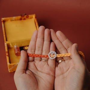 Puede incluir: Una pulsera trenzada con hilos naranjas y amarillos, con un dije plateado con el símbolo Om y pequeñas piedras transparentes. La pulsera se sostiene en manos abiertas, con una caja amarilla en el fondo.