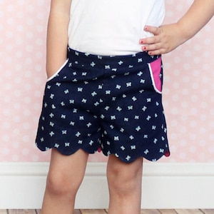 Puede incluir: Una niña pequeña con una camiseta blanca y pantalones cortos de estampado azul y blanco con ribete rosa. Los pantalones cortos tienen un dobladillo festoneado. La niña está de pie sobre un suelo de madera con un fondo de estampado rosa.