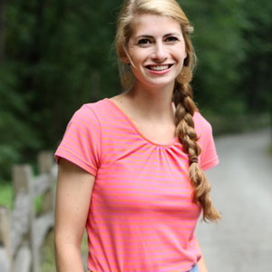 May include: A woman wearing a pink and orange striped short-sleeved shirt and a light blue skirt. She is holding a straw hat in her right hand.