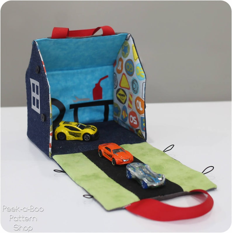 Peut inclure: Une maison de jeu en denim bleu et en tissu avec une poign&eacute;e rouge, ouverte pour r&eacute;v&eacute;ler une route verte et noire avec trois voitures miniatures. L'int&eacute;rieur de la maison de jeu est d&eacute;cor&eacute; d'un tissu bleu et blanc avec des panneaux de signalisation.