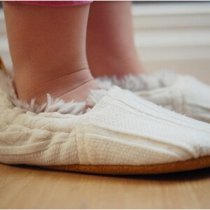 K&ouml;nnte beinhalten: Ein Paar wei&szlig;e gestrickte Babyschuhe mit flauschigem Besatz und einem gelben Akzent. Die Schuhe werden von einem Baby auf einem Holzboden getragen.