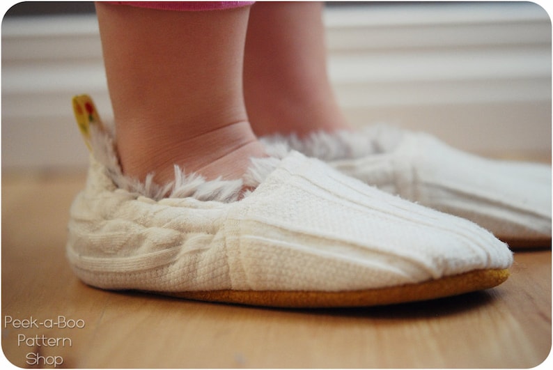 K&ouml;nnte beinhalten: Ein Paar wei&szlig;e gestrickte Babyschuhe mit flauschigem Besatz und einem gelben Akzent. Die Schuhe werden von einem Baby auf einem Holzboden getragen.