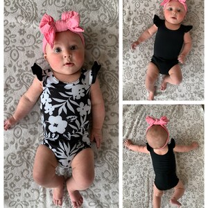May include: A baby wearing a black and white floral print onesie with ruffled sleeves and a pink bow headband. The baby is lying on a patterned blanket.