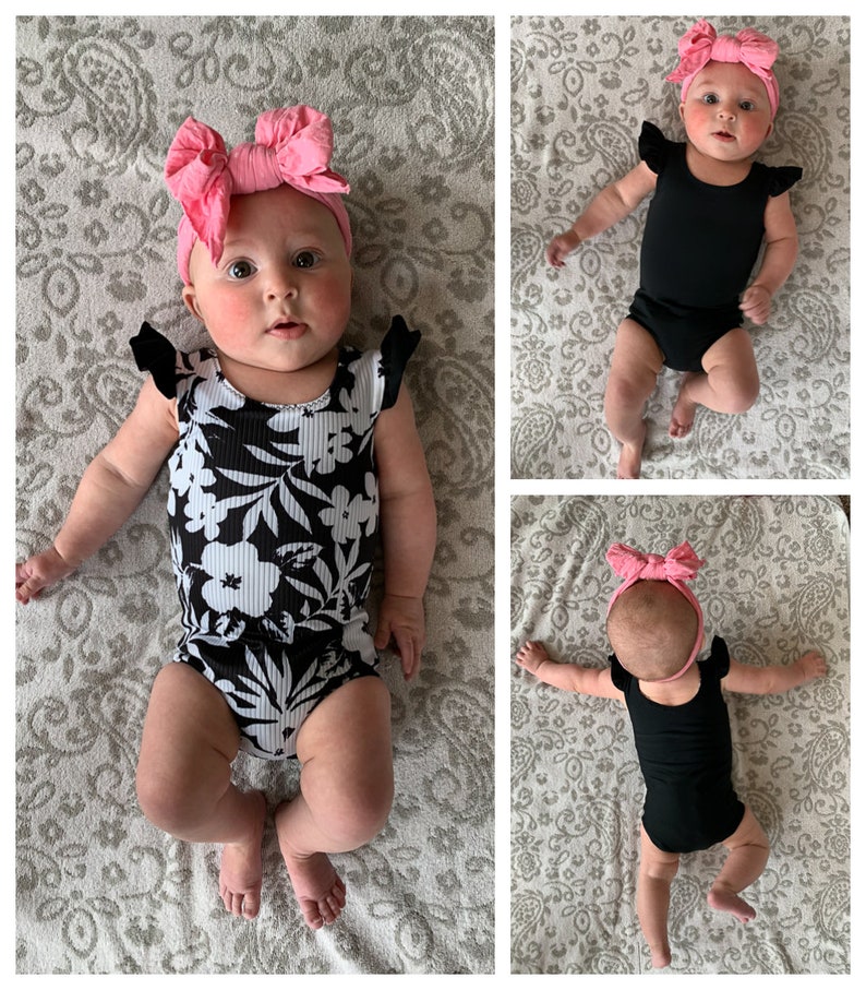 May include: A baby wearing a black and white floral print onesie with ruffled sleeves and a pink bow headband. The baby is lying on a patterned blanket.