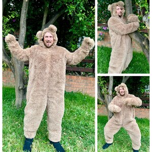May include: A person wearing a brown furry bear costume with a hood. The person is standing in a grassy area with trees in the background.