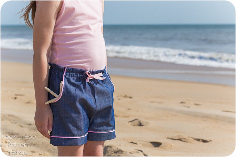 May include: A girl wearing a pink tank top and blue denim shorts with pink trim. The shorts have a drawstring waist and a starfish in the pocket. The girl is standing on a sandy beach with the ocean in the background.
