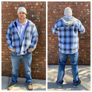 May include: A blue and black plaid flannel shirt with a gray hooded liner. The shirt is worn over a white collared shirt. The person is wearing a white beanie.