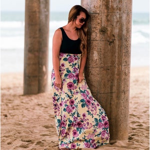May include: A woman wearing a black tank top and a yellow floral maxi skirt stands under a pier. The skirt has a purple floral pattern. The woman is leaning against a concrete pillar.