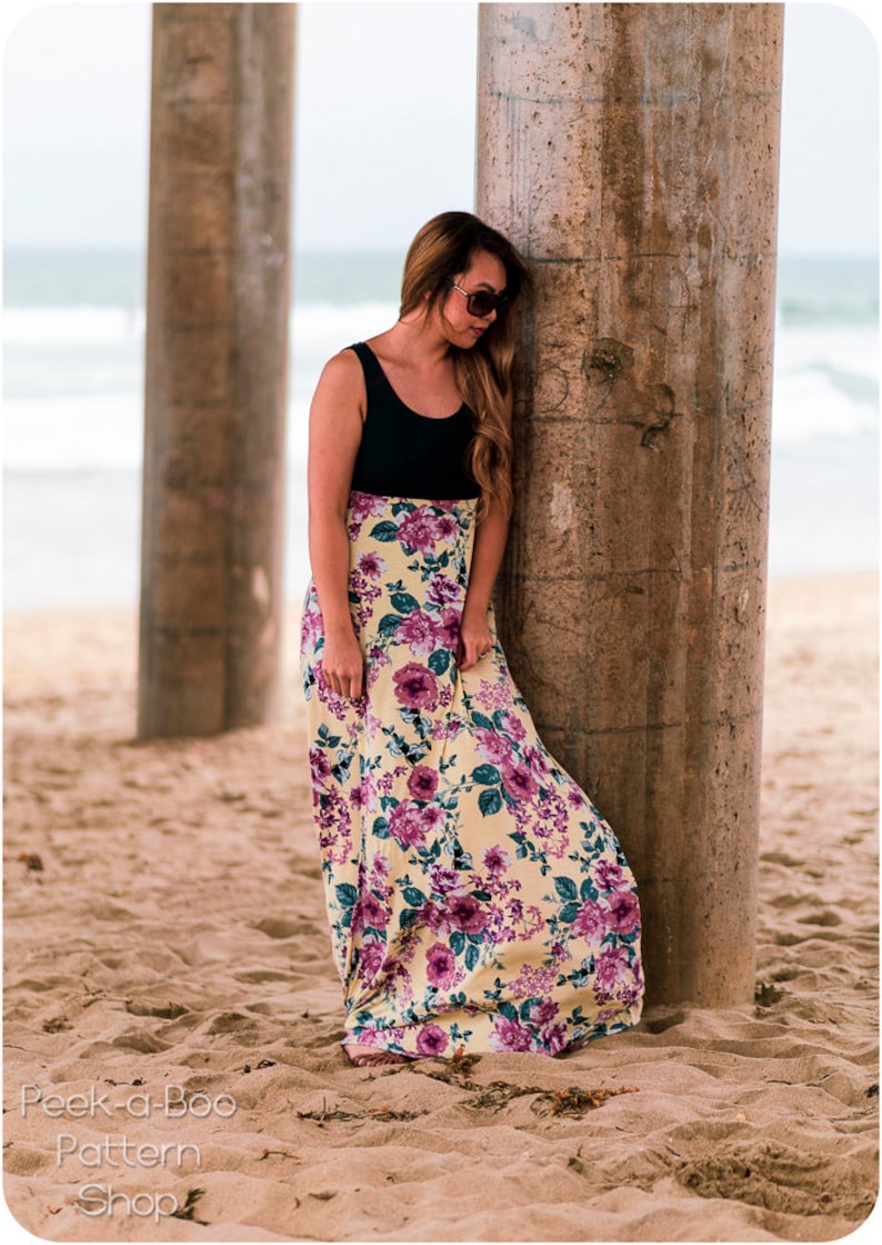 May include: A woman wearing a black tank top and a yellow floral maxi skirt stands under a pier. The skirt has a purple floral pattern. The woman is leaning against a concrete pillar.