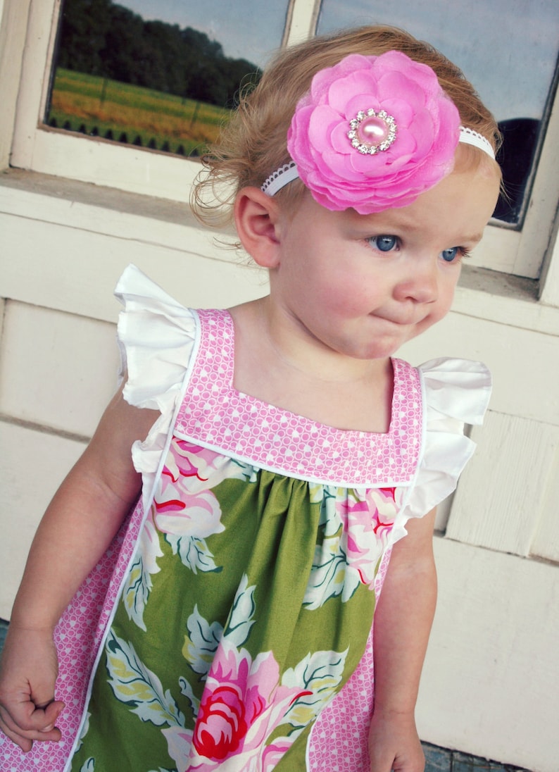 May include: A young girl wearing a green and pink floral dress with white ruffles on the sleeves. She is wearing a pink flower headband with a rhinestone in the center.