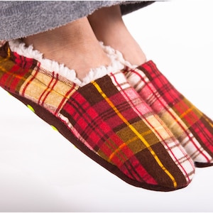 Può includere: Ciabatte a quadri rossi e marroni con dettagli bianchi e fodera bianca soffice. Le ciabatte sono indossate ai piedi di una persona.