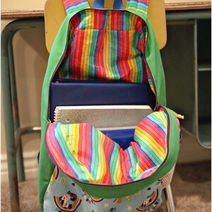 May include: A green and rainbow striped backpack with a blue zipper and a rainbow fabric lining. The backpack is open and filled with notebooks and a sparkly folder. The backpack is sitting on a chair.