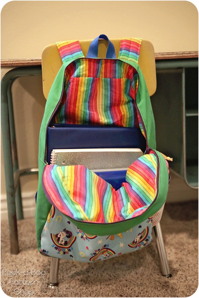 May include: A green and rainbow striped backpack with a blue zipper and a rainbow fabric lining. The backpack is open and filled with notebooks and a sparkly folder. The backpack is sitting on a chair.