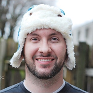 May include: A man wearing a white and blue winter hat with a faux fur brim. The hat has a flap that can be pulled down over the ears.
