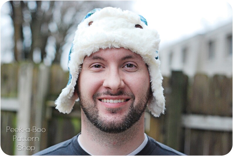 May include: A man wearing a white and blue winter hat with a faux fur brim. The hat has a flap that can be pulled down over the ears.