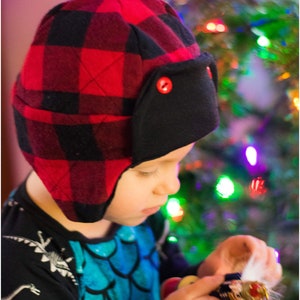 May include: A child wearing a red and black plaid hat with ear flaps and a blue and green shirt is holding a small ornament. The ornament is a figurine of a person with a white fur trim. The child is standing in front of a Christmas tree with colorful lights.