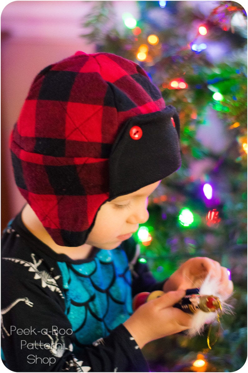 May include: A child wearing a red and black plaid hat with ear flaps and a blue and green shirt is holding a small ornament. The ornament is a figurine of a person with a white fur trim. The child is standing in front of a Christmas tree with colorful lights.